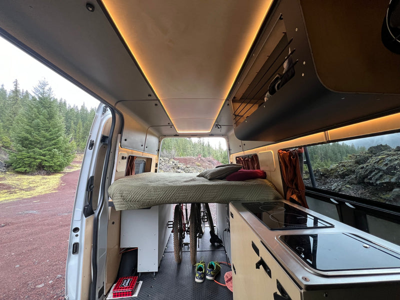 Inside a cozy RV rental: bed, kitchen, bikes below, with a scenic view of evergreen trees & rocks through the open rear. — photo 1