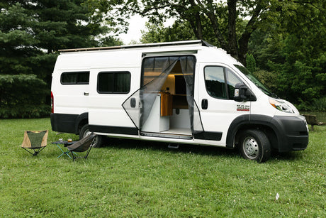 This white RV rental camper van is parked on lush grass, showcasing its open interior with a mesh screen and outdoor furniture.