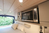 This RV rental interior features a Black+Decker microwave and control panel above a cozy bed, with a view of lush greenery.