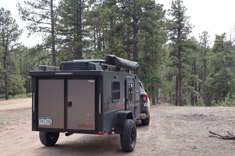 Rugged inTech Flyer adventure trailer (RV rental) with solar and awning, parked in a forest campsite.