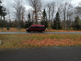 Autumn RV rental: A red camper van is parked on a path in a park, with a person nearby and fall leaves covering the ground under a cloudy sky.