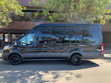 Sleek black Ford Transit conversion van, an ideal rv rental, parked on a sunlit street reflecting trees & architecture.