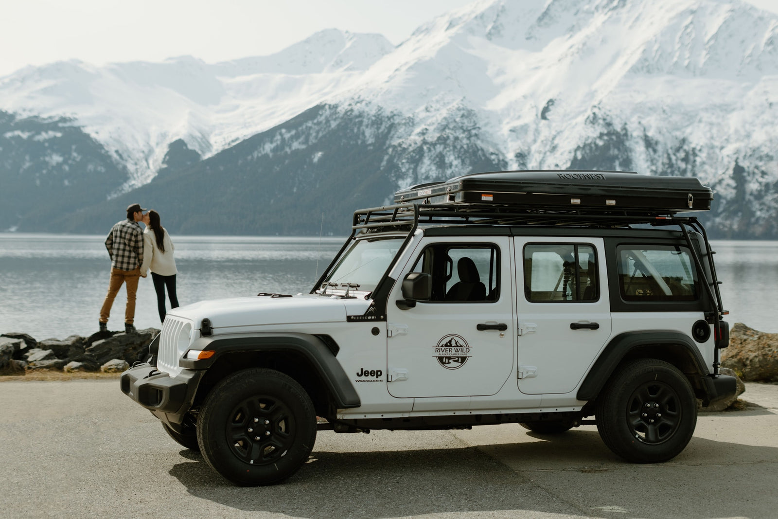 White Jeep RV rental with rooftop tent by an Alaskan lake. Couple enjoys snowy mountain views.