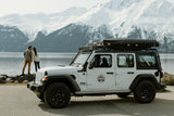 White Jeep RV rental with rooftop tent by an Alaskan lake. Couple enjoys snowy mountain views.