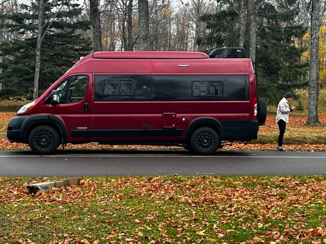  ideal for RV rental getaways. Fall trees frame this adventure vehicle.|A striking red **rv rental** camper van parked roadside amidst vibrant autumn leaves. A person checks their phone by the back.|Maroon off-road RV rental parked on a wet driveway — photo 2