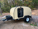 Tan teardrop camper trailer parked on a paved driveway, surrounded by trees. Ideal RV rental for outdoor trips.