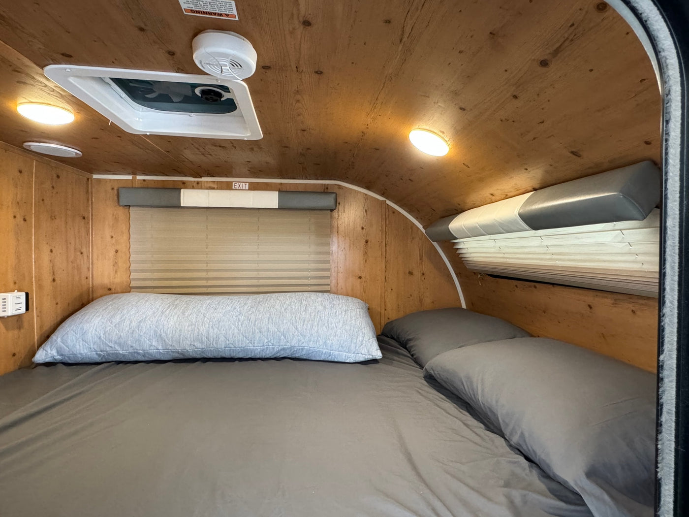 Comfortable RV rental interior showing a bed with grey bedding, wood walls, ceiling fan, and window blinds. — photo 2
