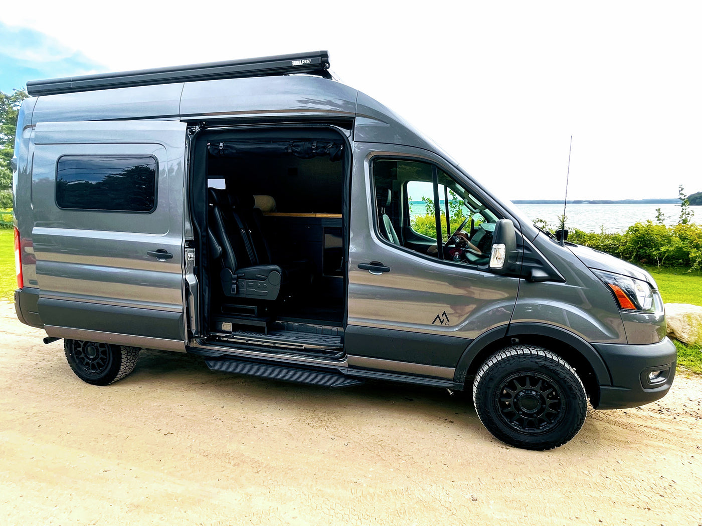 Explore with this grey camper van RV rental parked by a lake. Its side door is open, revealing a custom interior.