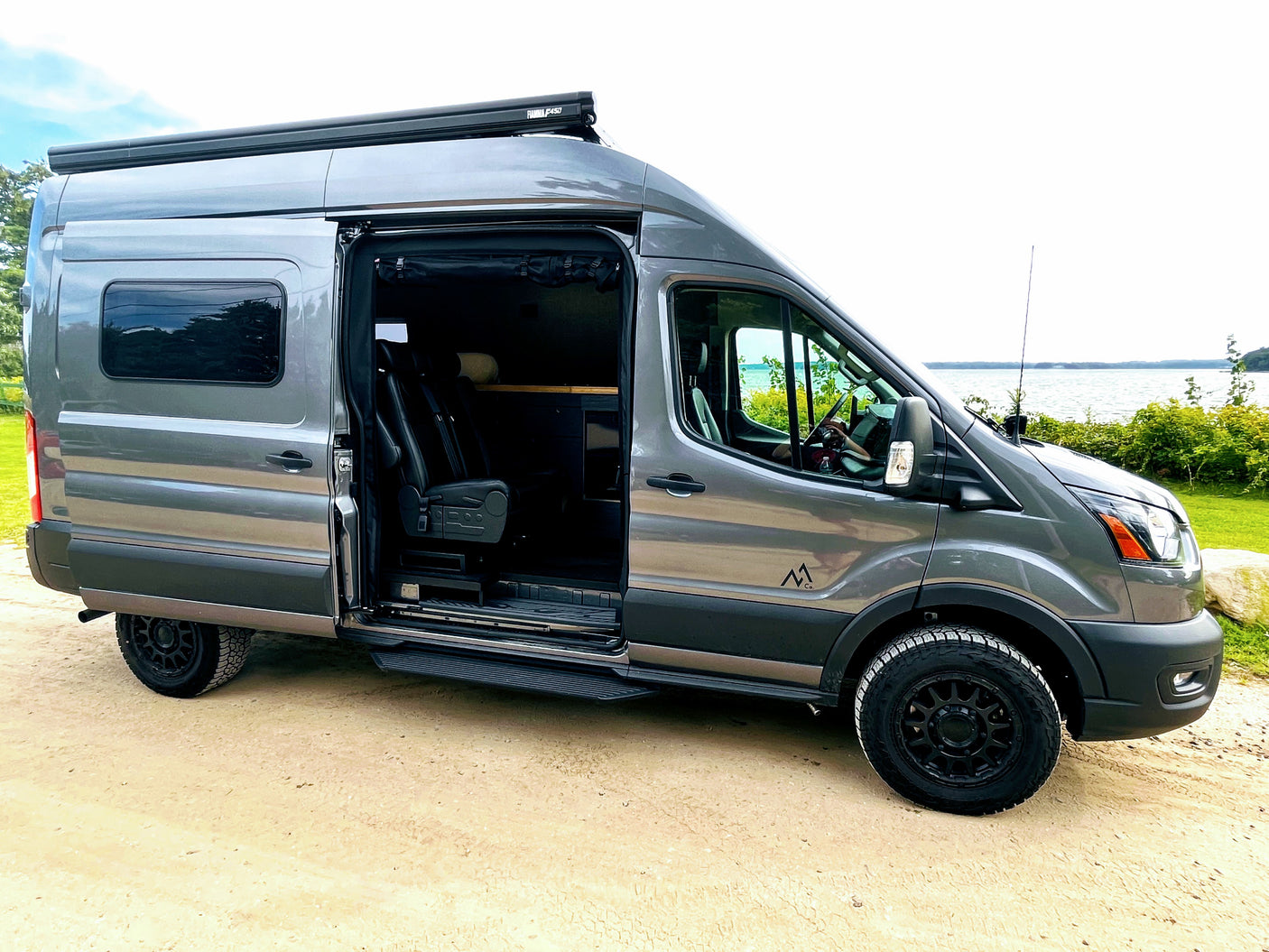 Explore with this grey camper van RV rental parked by a lake. Its side door is open, revealing a custom interior.