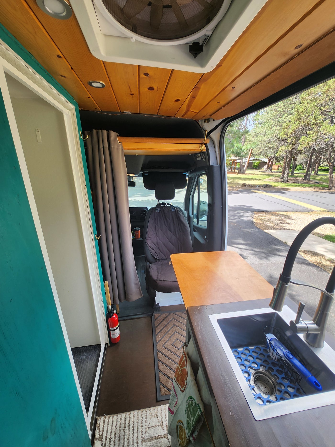 RV rental camper van interior. Wood ceiling, modern sink/faucet, teal partition, and driver's area view. Essentials include a fire extinguisher. — photo 7