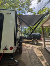 White rv rental camper with its awning extended, providing shade over a black SUV at a sunny, tree-lined campsite.