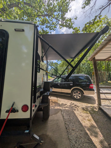 White rv rental camper with its awning extended, providing shade over a black SUV at a sunny, tree-lined campsite.