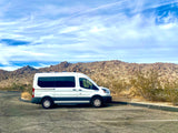 White RV rental van parked in a desert landscape with rocky hills under a blue sky.