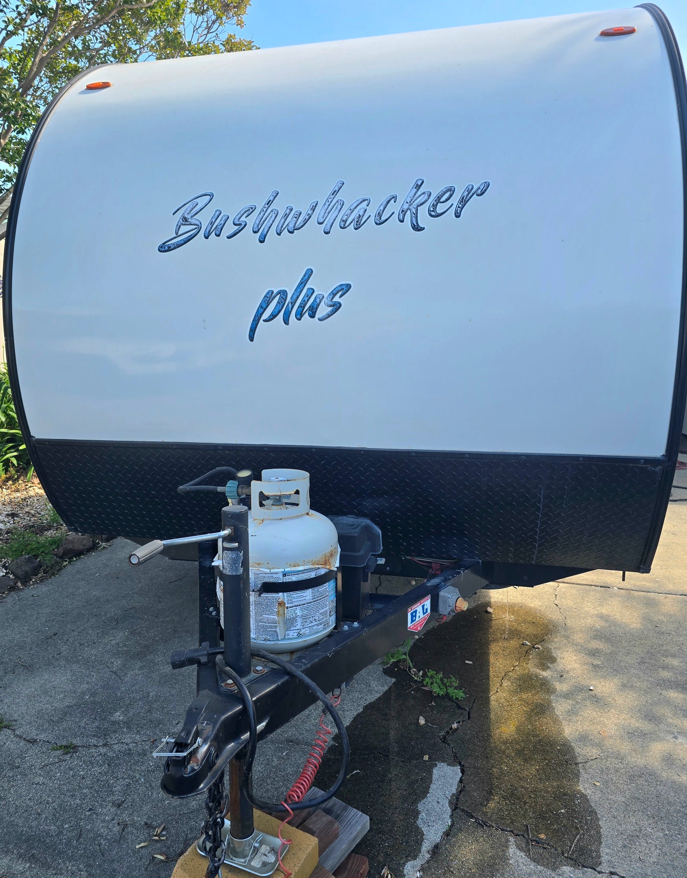 Bushwhacker Plus rv rental: Front of a white travel trailer with a propane tank and hitch assembly, parked on concrete.