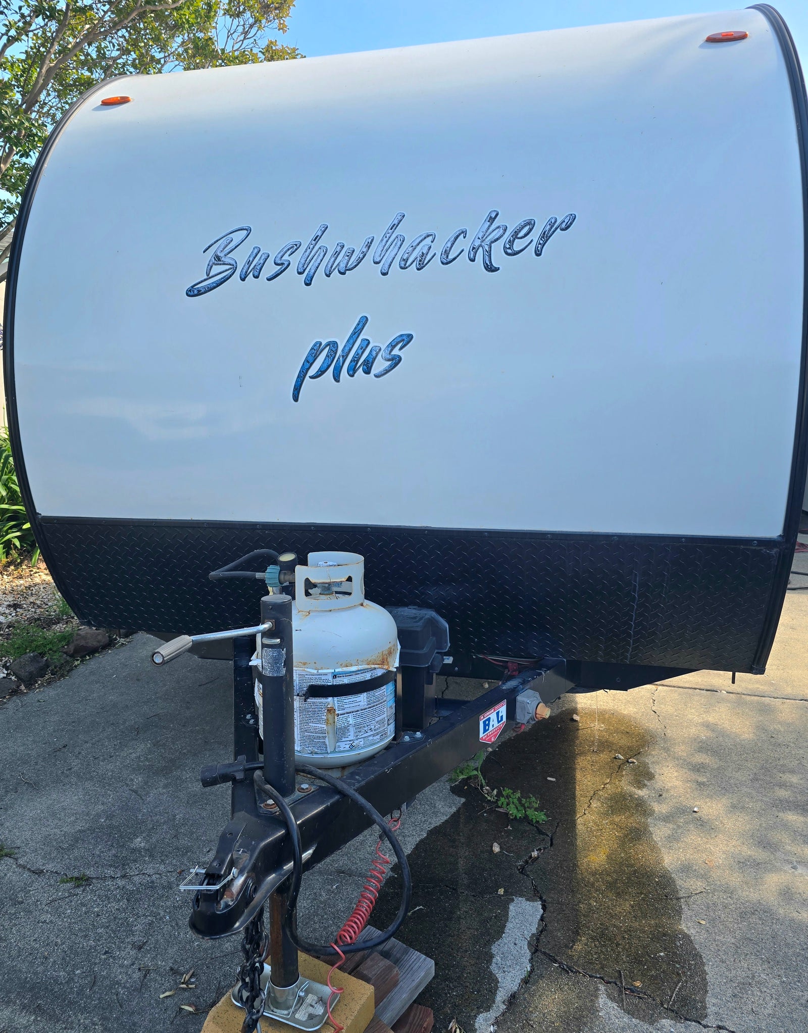 Bushwhacker Plus rv rental: Front of a white travel trailer with a propane tank and hitch assembly, parked on concrete.
