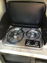 An RV rental features a compact FlameKing two-burner stove with a protective wind guard, ideal for camping.