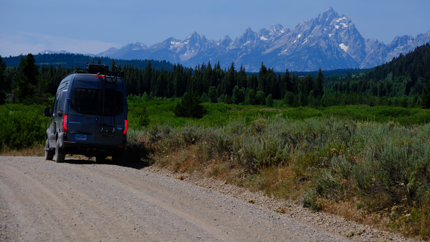 An RV rental camper van on a scenic dirt road, backed by the impressive Teton mountains and lush green forests.