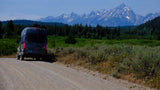 An RV rental camper van on a scenic dirt road, backed by the impressive Teton mountains and lush green forests.
