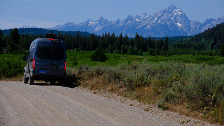 An RV rental camper van on a scenic dirt road, backed by the impressive Teton mountains and lush green forests.