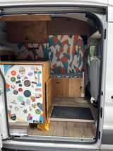 RV rental: Custom camper van interior with wood cabinetry, a colorful geometric wall, and a unique sticker collection.
