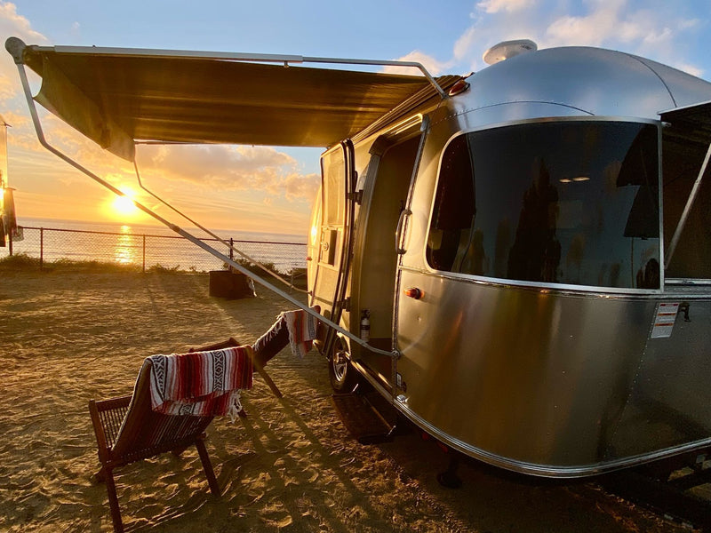  complete with a cozy chair under the awning.|Shiny Airstream RV rental parked under large trees at a campground — photo 1