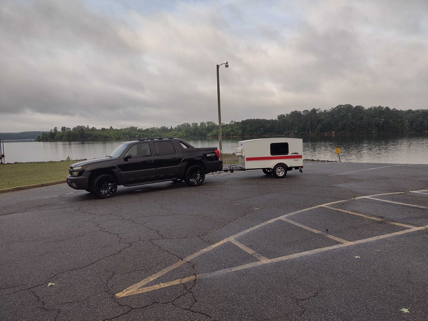 Black truck with a compact RV rental (teardrop camper) at a boat launch by a lake on a cloudy day.