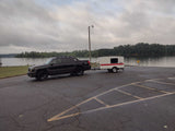 Black truck with a compact RV rental (teardrop camper) at a boat launch by a lake on a cloudy day.