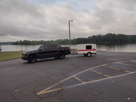 Black truck with a compact RV rental (teardrop camper) at a boat launch by a lake on a cloudy day.