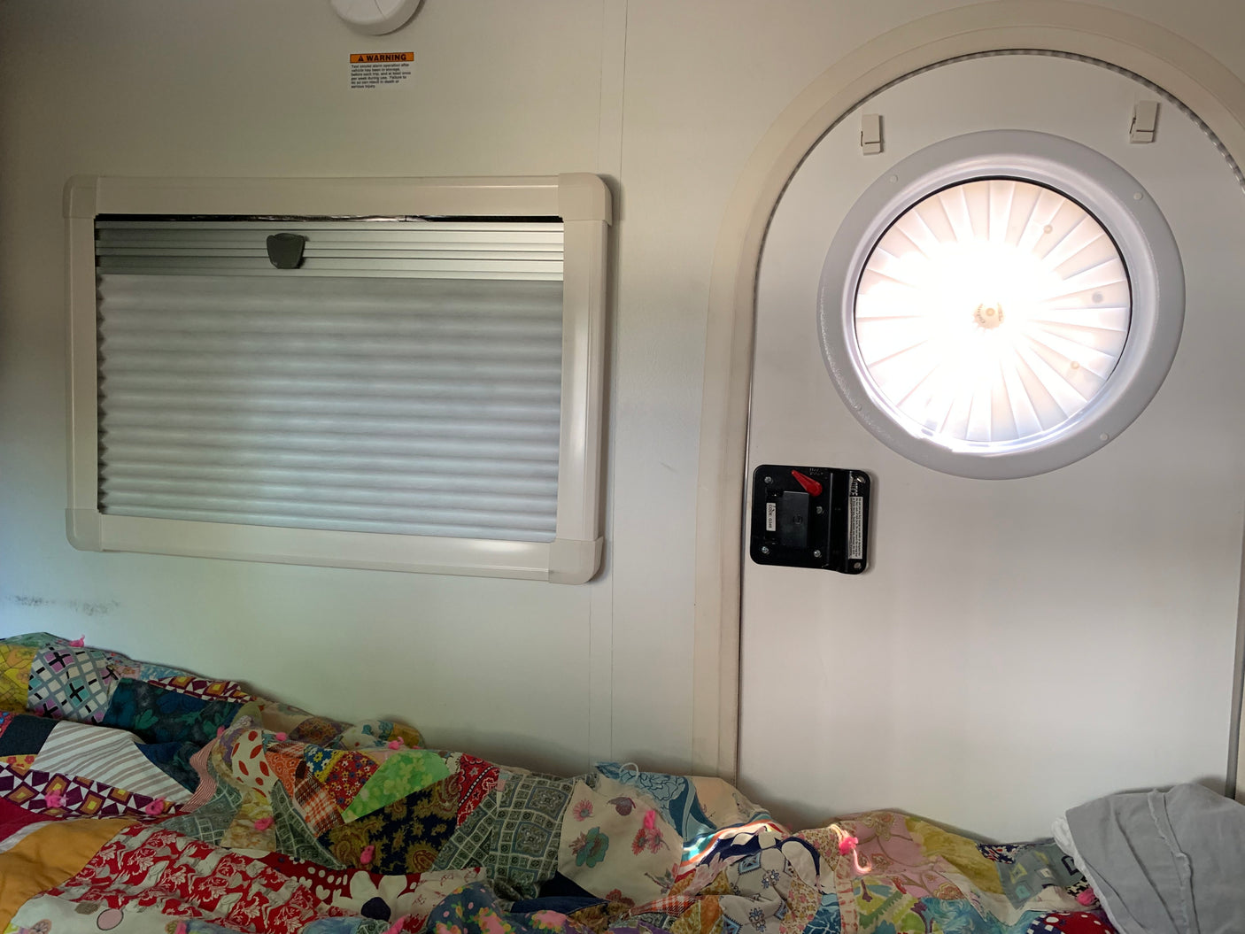 Cozy RV rental interior shows a bright circular door, a window with pleated blinds, and a colorful patchwork quilt on the bed.