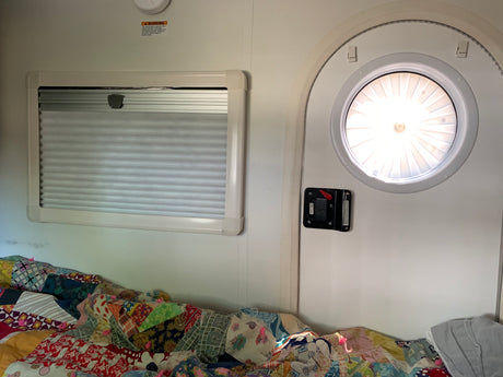 Cozy RV rental interior shows a bright circular door, a window with pleated blinds, and a colorful patchwork quilt on the bed.