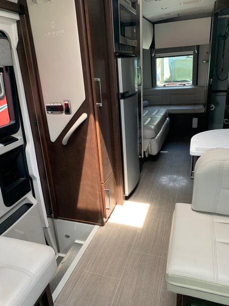 Modern rv rental interior: light leather seating, wood cabinetry, built-in kitchen (microwave, fridge), and an aisle leading to rear bench.