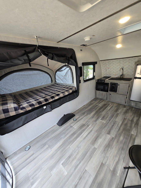Compact RV rental interior: pop-out bed with plaid blanket, kitchenette with sink & stove, and wood-look flooring.
