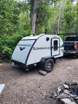 Bushwhacker travel trailer, perfect for an rv rental, parked at a leafy campsite next to a pickup truck and fire pit.