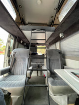 RV rental interior: Functional camper van showing front seats, a ladder to an upper bunk bed, and a rear bench seat.