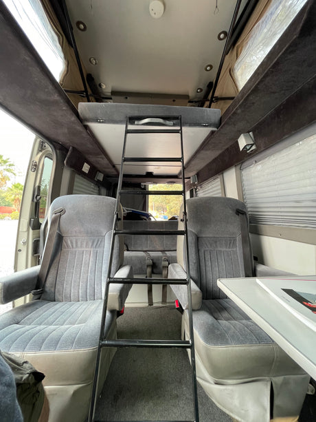 RV rental interior: Functional camper van showing front seats, a ladder to an upper bunk bed, and a rear bench seat.