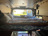 Cozy bed in an RV rental, featuring patterned blankets and pillows, with a bright view of a yard through the window.