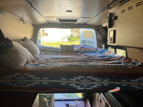 Cozy bed in an RV rental, featuring patterned blankets and pillows, with a bright view of a yard through the window.