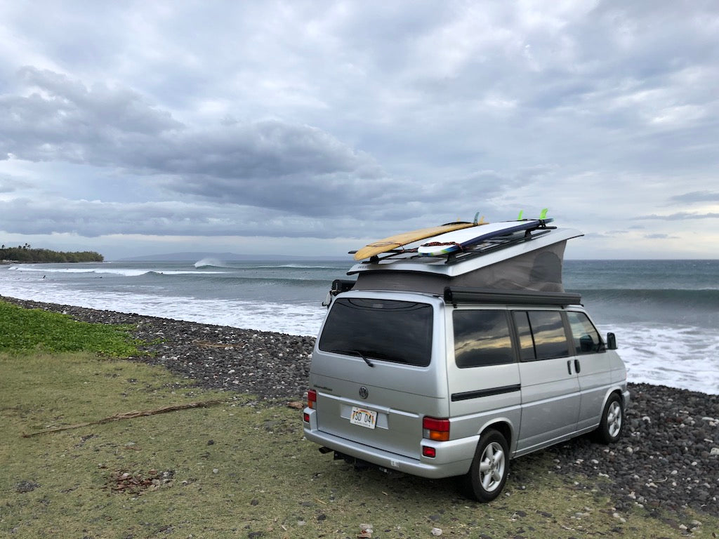 Silver RV rental van with surfboards on roof, parked at a rocky beach with waves and distant surfers under cloudy skies. — photo 8