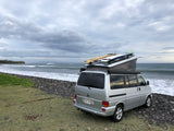 Silver RV rental van with surfboards on roof, parked at a rocky beach with waves and distant surfers under cloudy skies.