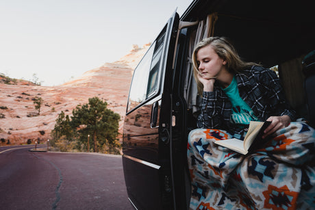 Relaxing RV rental moment: Young woman reading in van door, cozy in a blanket, overlooking scenic desert.