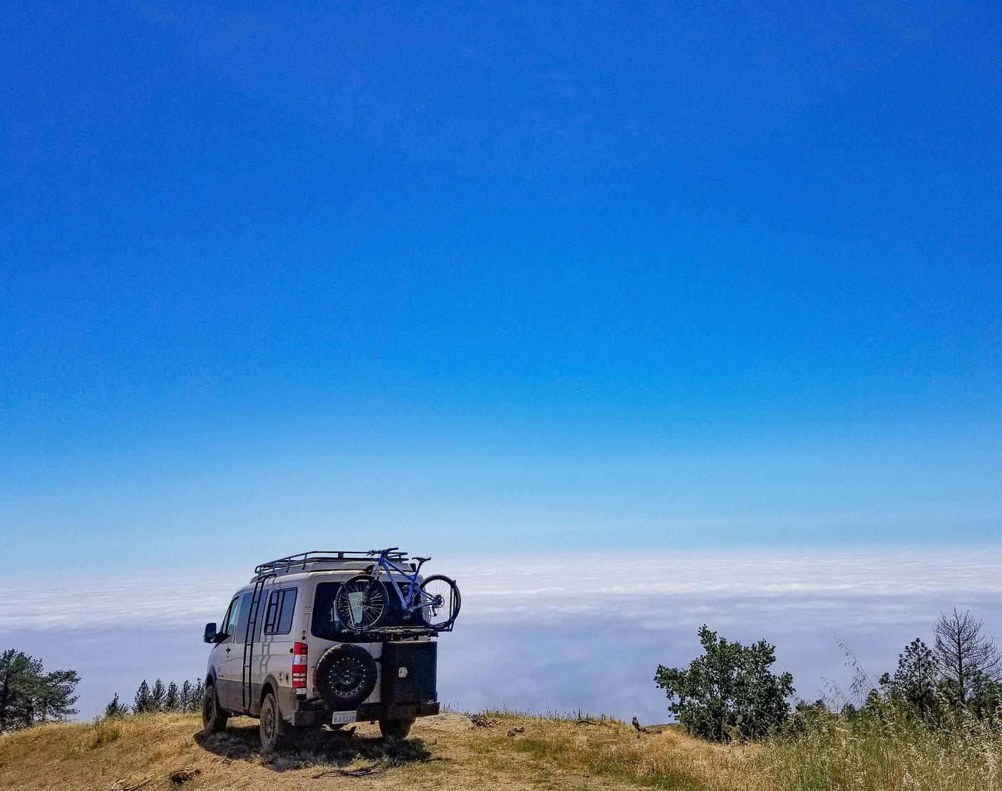 RV rental van with bike parked on a mountain, overlooking a vast sea of clouds under a clear blue sky. Adventure awaits!