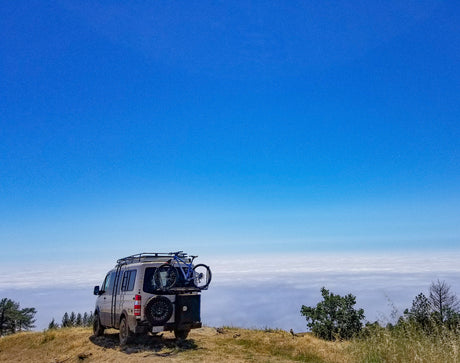 RV rental van with bike parked on a mountain, overlooking a vast sea of clouds under a clear blue sky. Adventure awaits!