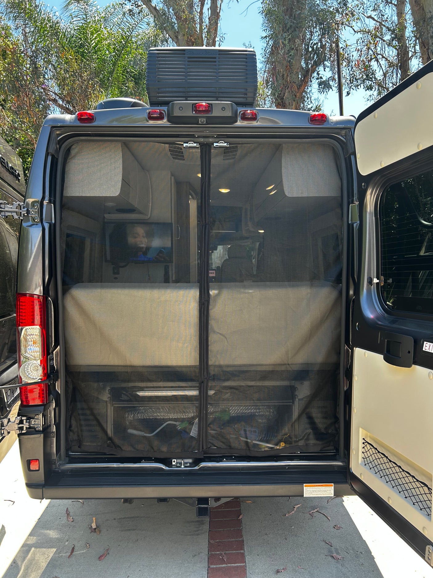 Rear view of an RV rental with open doors and a mesh screen, revealing a cozy interior with a bed and TV.