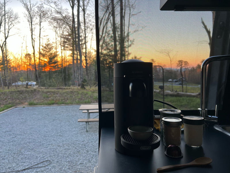 Inside an RV rental, a Nespresso machine and coffee supplies sit on a counter, with a vibrant sunset over a wooded campsite visible through the screened window. — photo 6