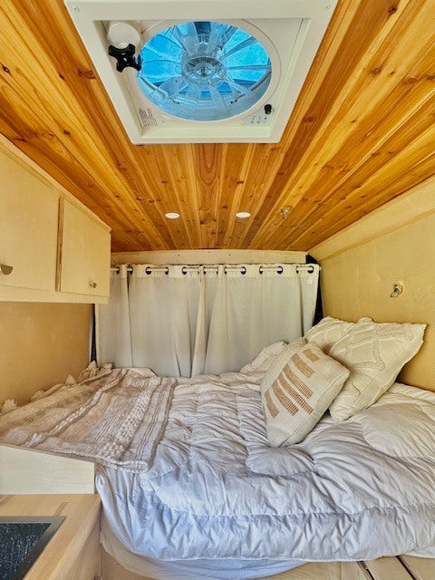 Cozy RV rental interior showing a comfortable bed, wood-paneled ceiling with a fan, and white curtains.