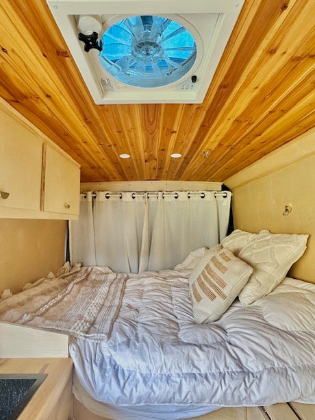 Cozy RV rental interior showing a comfortable bed, wood-paneled ceiling with a fan, and white curtains.