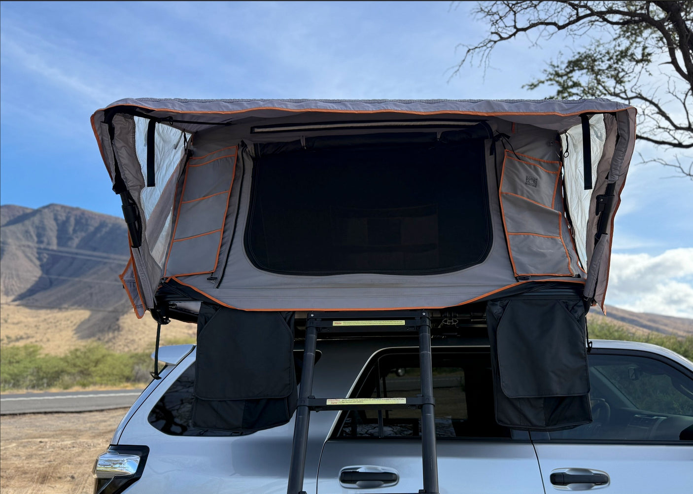This RV rental SUV features an open grey rooftop tent with interior mesh, storage pockets, and a ladder, ready for outdoor adventure against a mountain backdrop.