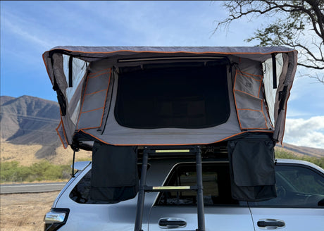This RV rental SUV features an open grey rooftop tent with interior mesh, storage pockets, and a ladder, ready for outdoor adventure against a mountain backdrop.