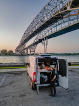 Two people enjoy the scenic river view from their RV rental van, parked beneath a majestic bridge at sunset.