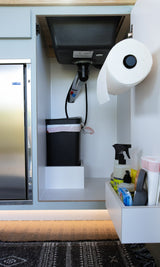 Under-sink area in an RV rental, showing a trash can, paper towels, cleaning supplies, and a compact fridge. Optimized storage.